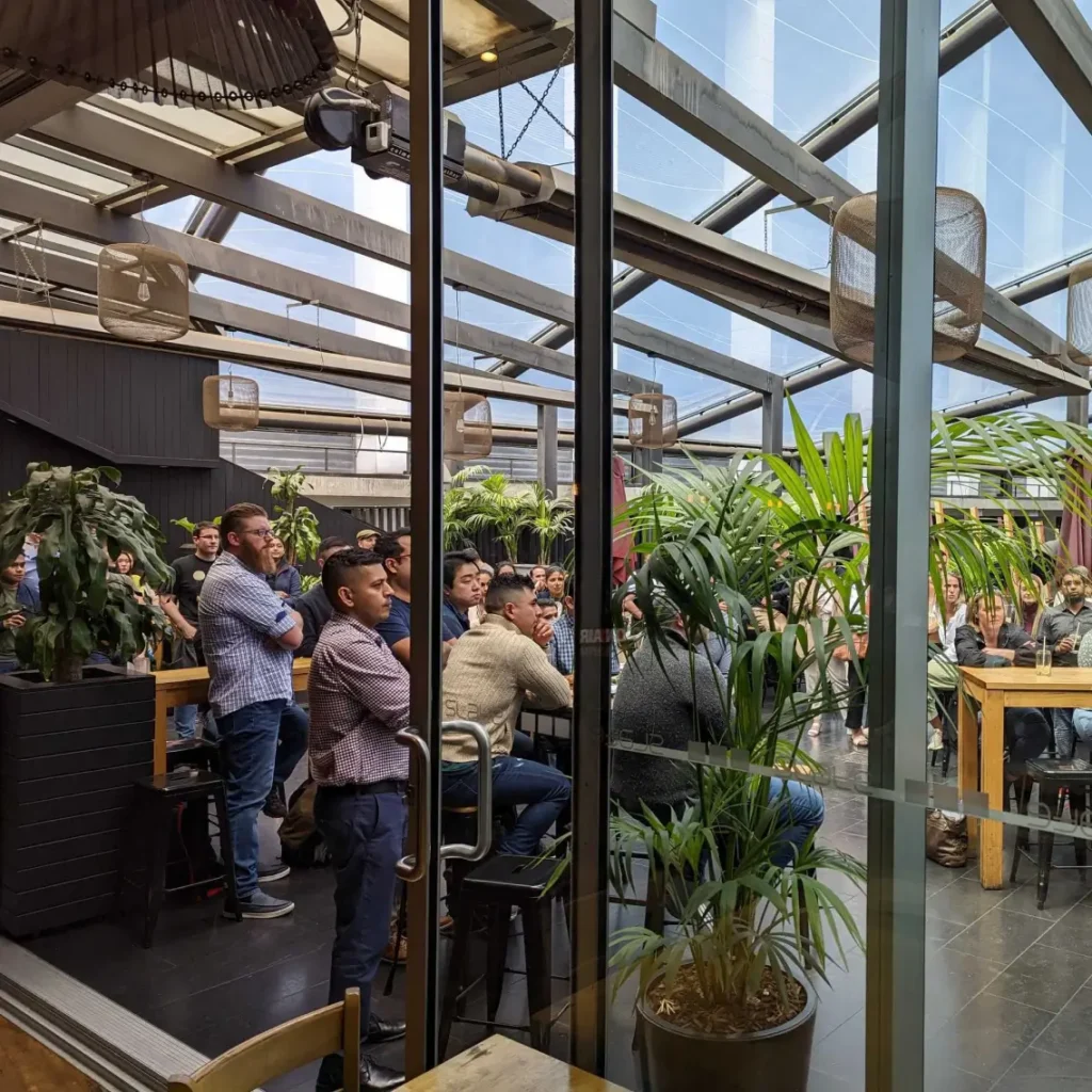A group of people sit and stand attentively in a modern, glass-roofed indoor space with large windows, plants, and wooden tables, suggesting a meeting or presentation is taking place.