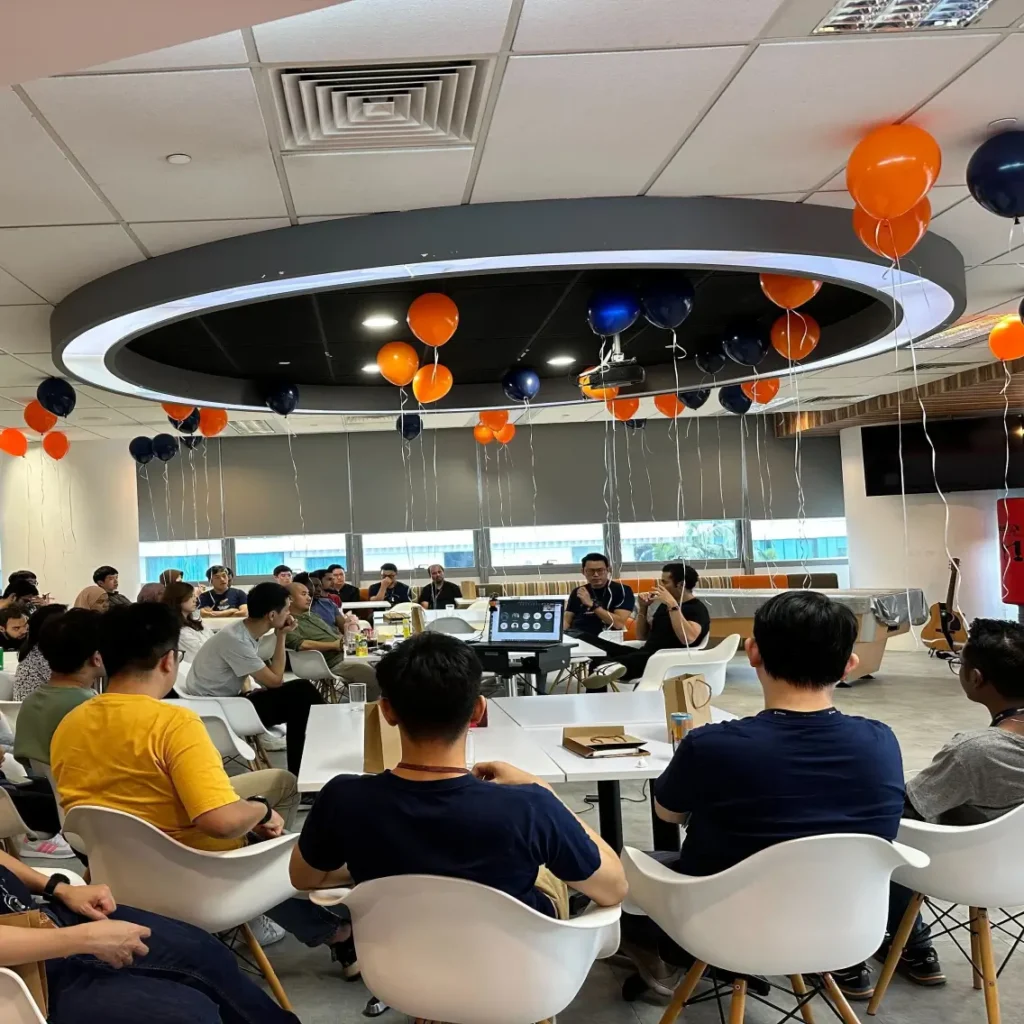A group of people sits around tables in a modern office space decorated with orange and dark blue balloons hanging from the ceiling, attending a meeting or presentation.