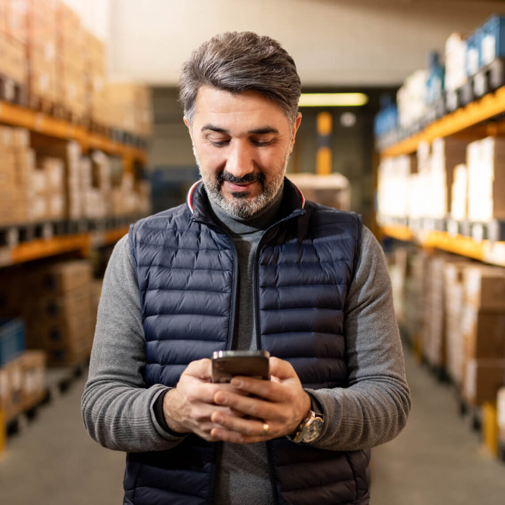 A man with gray hair and a beard, wearing a gray turtleneck and navy puffer vest, stands in a warehouse aisle smiling while looking at his smartphone. Shelves with boxes are visible in the background.