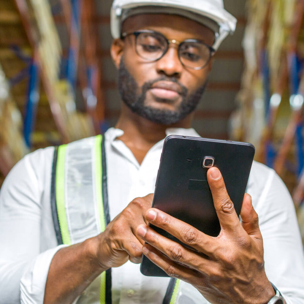 A man wearing a hard hat, safety vest, and glasses is standing indoors, using a tablet. The background is blurred, suggesting a warehouse or industrial setting.