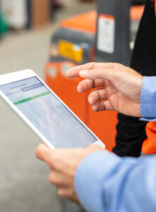 Close-up of a person using a tablet, displaying a spreadsheet or data, with another person nearby. An orange vehicle or machinery is blurred in the background.