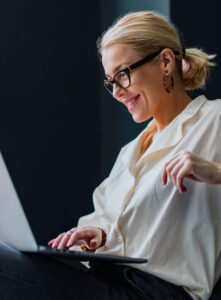A woman with blonde hair in a ponytail, wearing glasses and a white shirt, smiles as she uses a laptop. She is sitting indoors against a dark background.