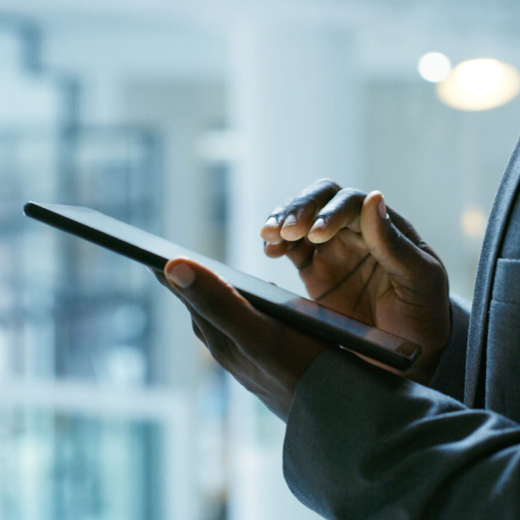 A person in a suit is using a touchscreen tablet, with one hand holding the device and the other hand tapping the screen. The background is softly blurred, suggesting an indoor setting, possibly an office.