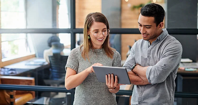 Two people standing in an office, smiling and looking at a tablet together. The woman is holding the tablet and pointing at the screen, while the man stands beside her with his arms crossed.