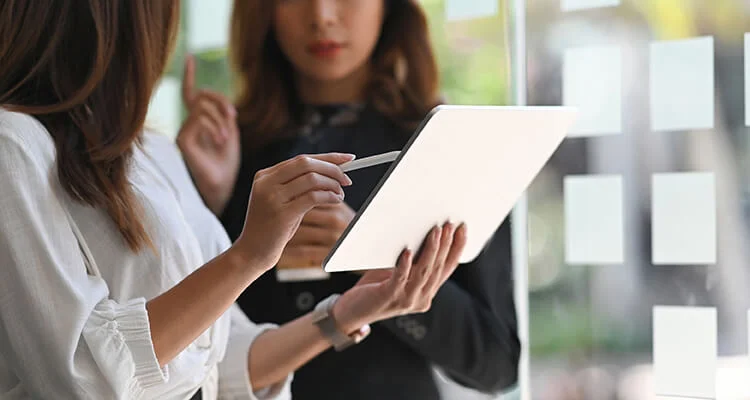 Two women stand indoors; one holds a digital tablet while the other gestures with a finger. Both are focused on the screen, appearing engaged in discussion or collaboration.