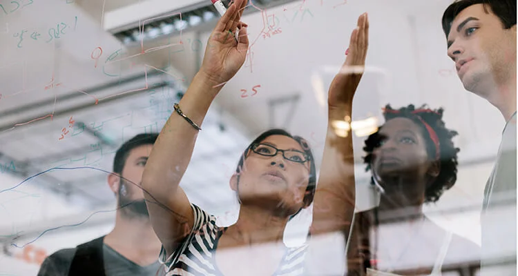 Four people stand behind a transparent board covered in graphs and equations; one person gestures towards the board, explaining something to the group.