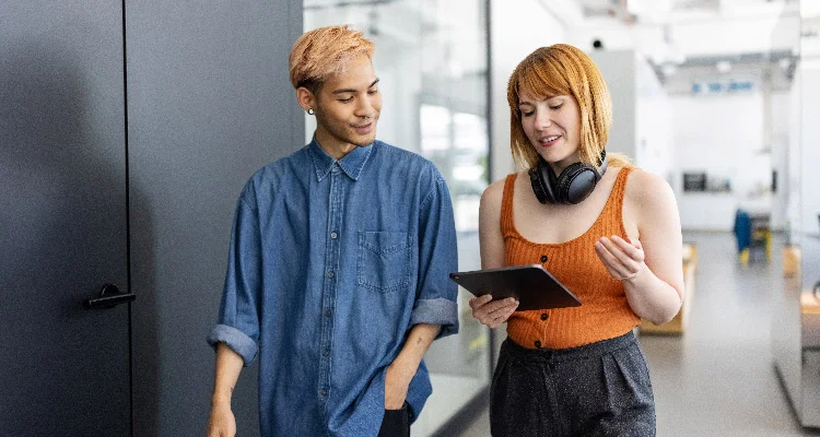 Two people walk together indoors. One is holding a tablet and wearing headphones around their neck, while the other smiles and looks at the tablet. They appear to be having a casual conversation in a modern office setting.