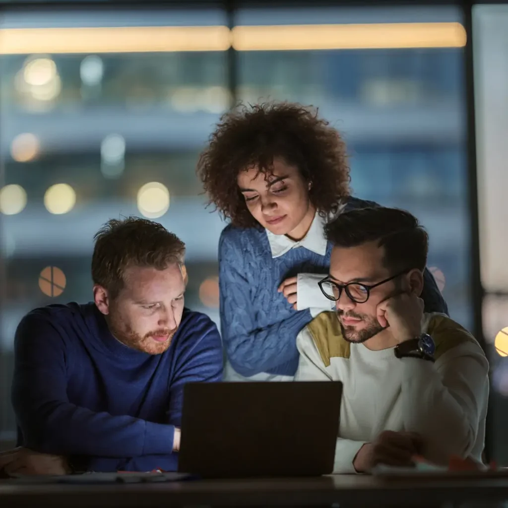 Three people are gathered around a laptop in an office at night, focused and working together. The background shows blurred city lights through large windows.