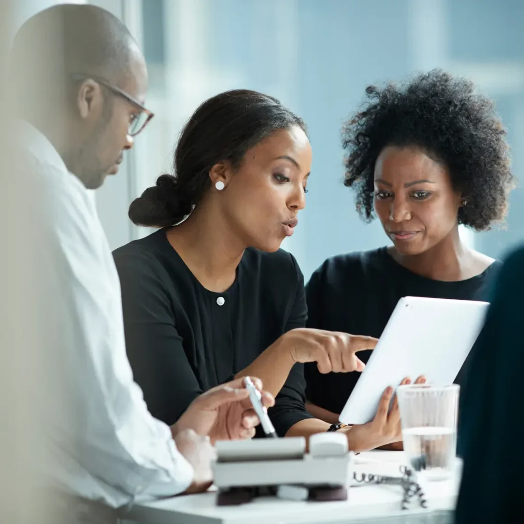 Three professionals in business attire have a discussion around a table. Two women are looking at and interacting with a tablet, while a man listens attentively. The setting appears to be a bright, modern office.