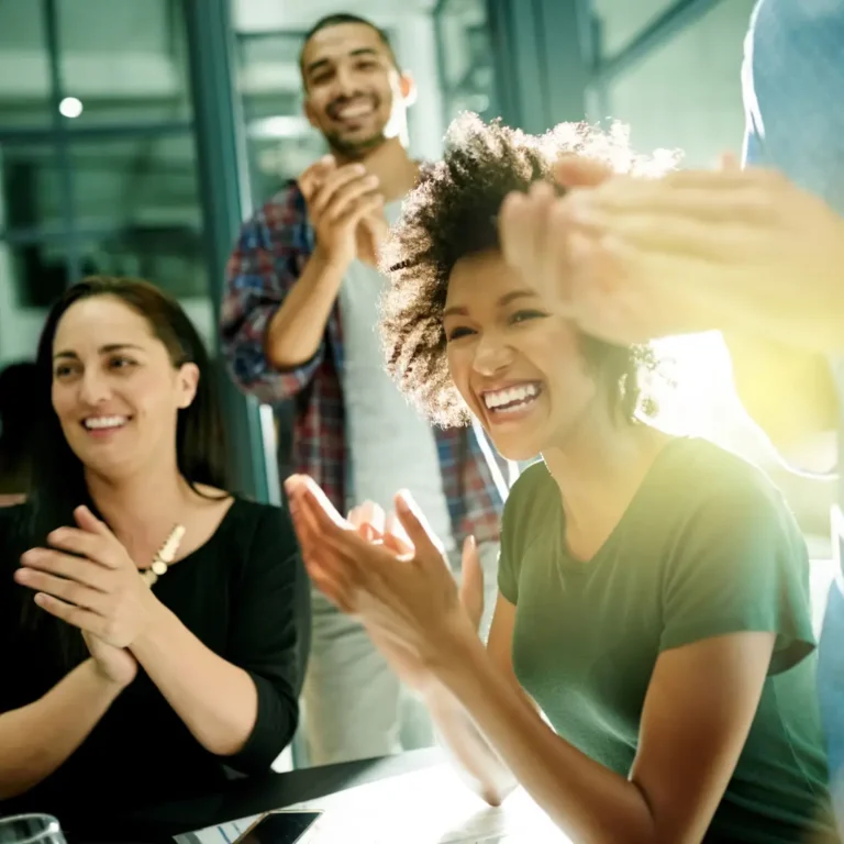 Four people in a bright office setting are smiling and clapping, celebrating or applauding something together. Sunlight streams in from a window, highlighting their joyful expressions.