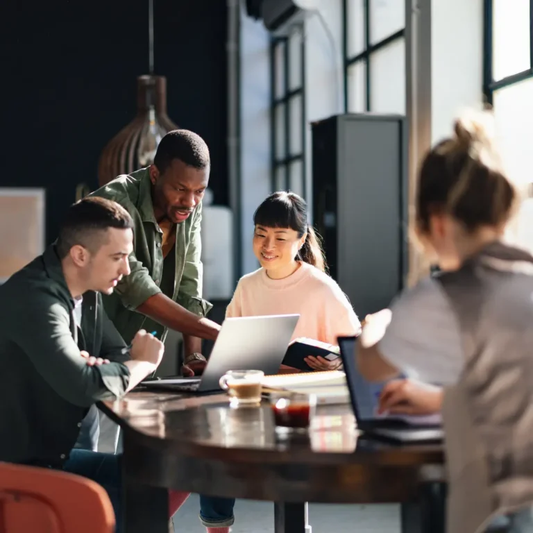 Three people sit and one stands at a table in a bright office, collaborating and looking at a laptop. Another person works on a laptop in the foreground, slightly out of focus.