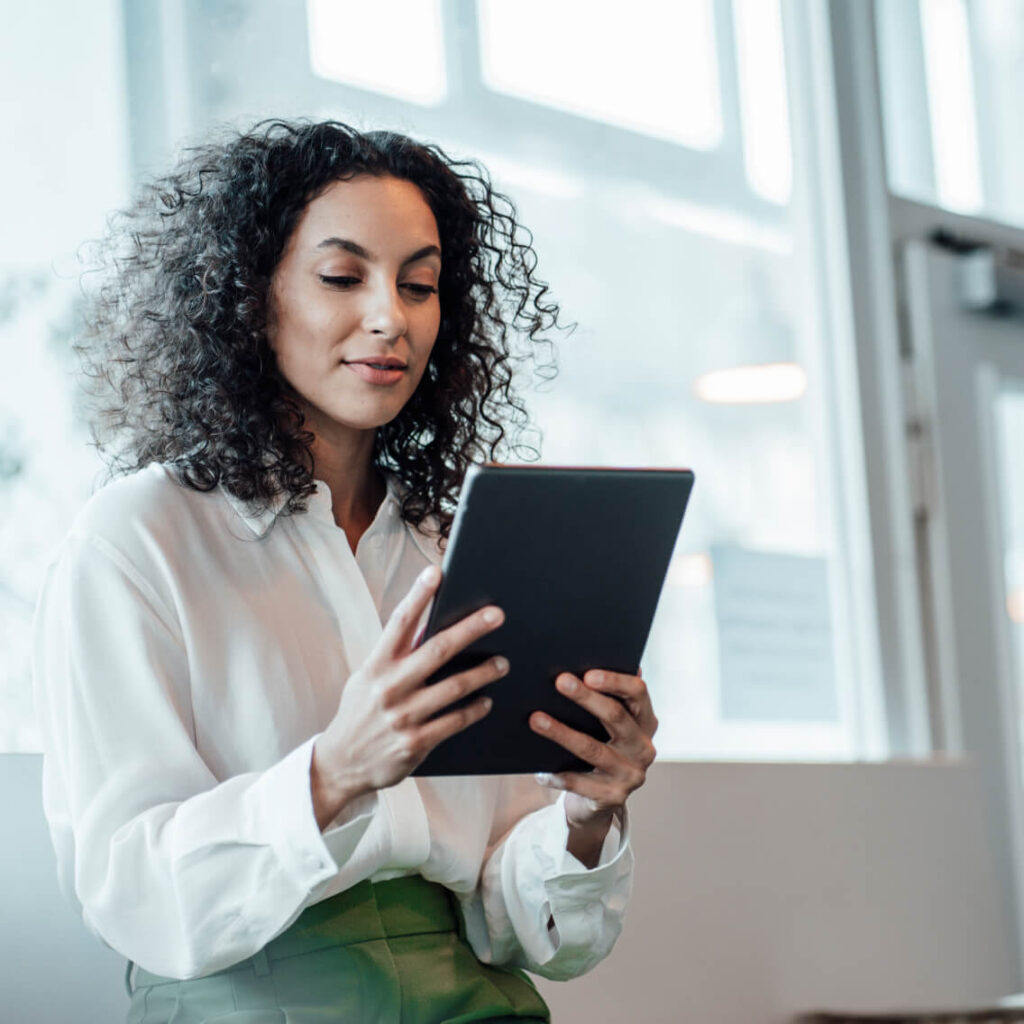 A woman with curly hair wearing a white blouse reads or works on a tablet in a bright indoor setting with large windows in the background.