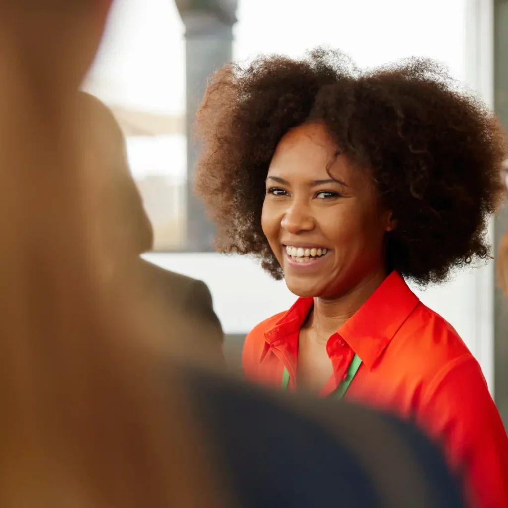 A woman with curly hair wearing a bright orange shirt smiles warmly while interacting with others in a well-lit indoor setting.