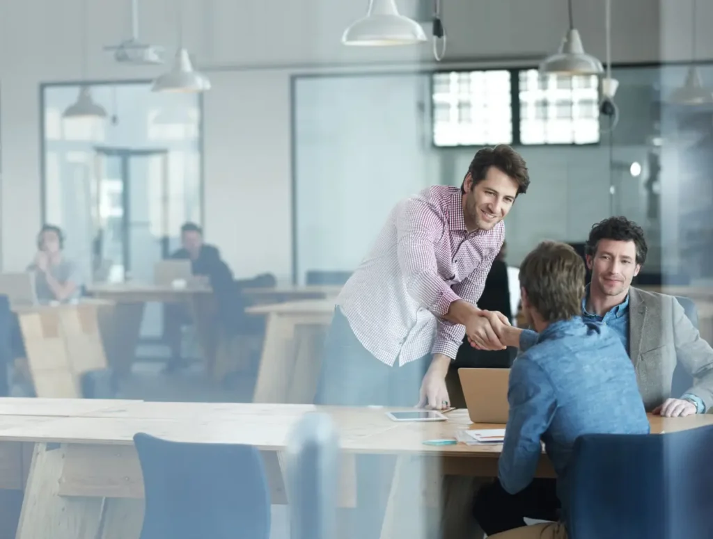 Three men are sitting at a shared table in a modern office. One man is standing and leaning forward to shake hands with another, while the third man sits nearby. Other people work in the background.