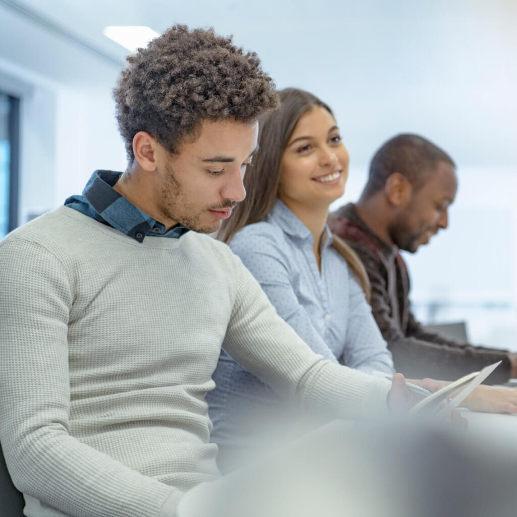 Three young adults sit side by side at a desk; one is writing, one is smiling at the camera, and one is focused on a tablet. The setting appears to be a bright, modern classroom or office.