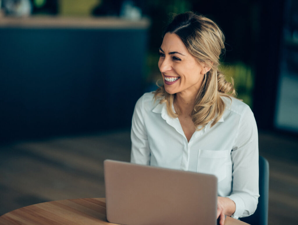 A woman with blonde hair, wearing a white shirt, sits at a table with a laptop and smiles while looking to the side in a bright, modern indoor setting.