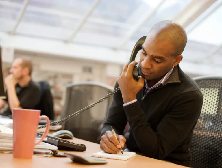 A man sits at a desk in an office, holding a phone between his ear and shoulder while writing in a notebook. A pink mug, papers, and a calculator are on the desk. Another person sits blurred in the background.