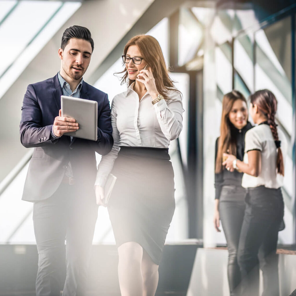Two business professionals, a man and a woman, walk together in an office corridor. The man holds a tablet and the woman is smiling, holding a phone. Two other women are talking in the blurred background.