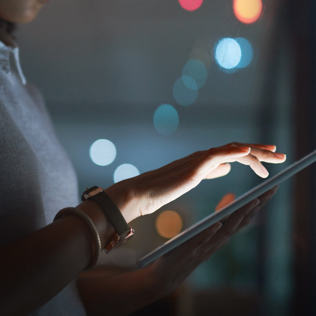 Close-up of a person’s hands using a touchscreen tablet in a dimly lit setting with blue and red bokeh lights in the background. The person is wearing a gray shirt and a wristwatch.