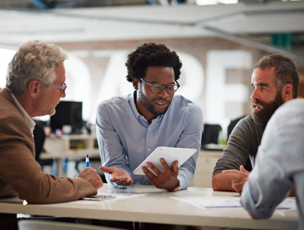Three men sitting at a table in an office, having a discussion. One man in the center is holding a tablet and talking, while the other two listen attentively, with documents and pens on the table.