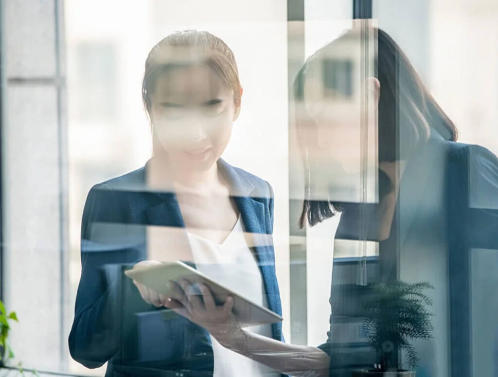 Two women in business attire stand by a window, looking at a tablet together. The image is partly obscured by reflections on the glass, giving a layered and slightly blurred effect.