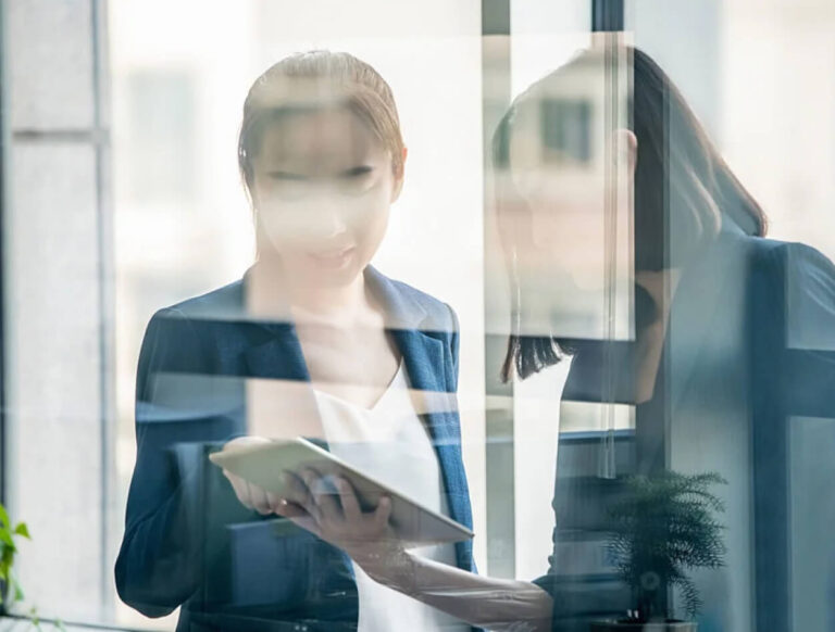 Two women in business attire stand by a window, looking at a tablet together. The image is partly obscured by reflections on the glass, giving a layered and slightly blurred effect.