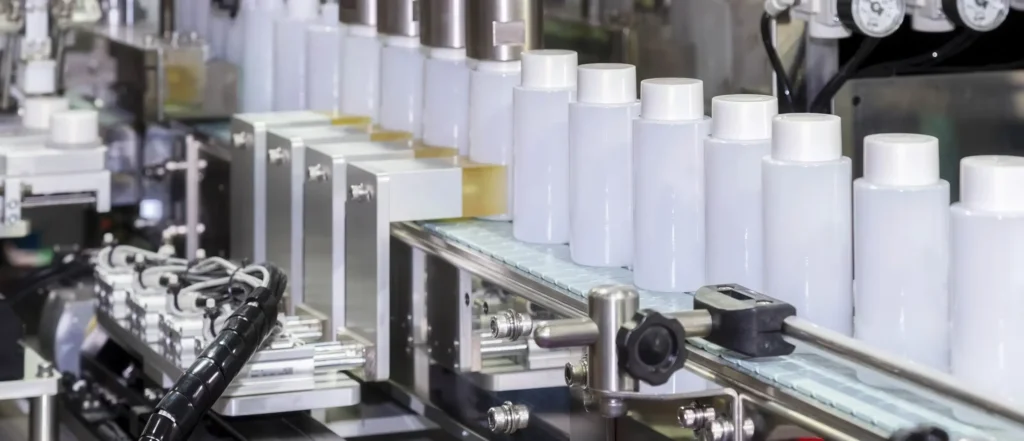 White plastic bottles move along a conveyor belt in a factory setting, likely part of an automated production or packaging line for liquid products. Machinery and metal components surround the bottles.