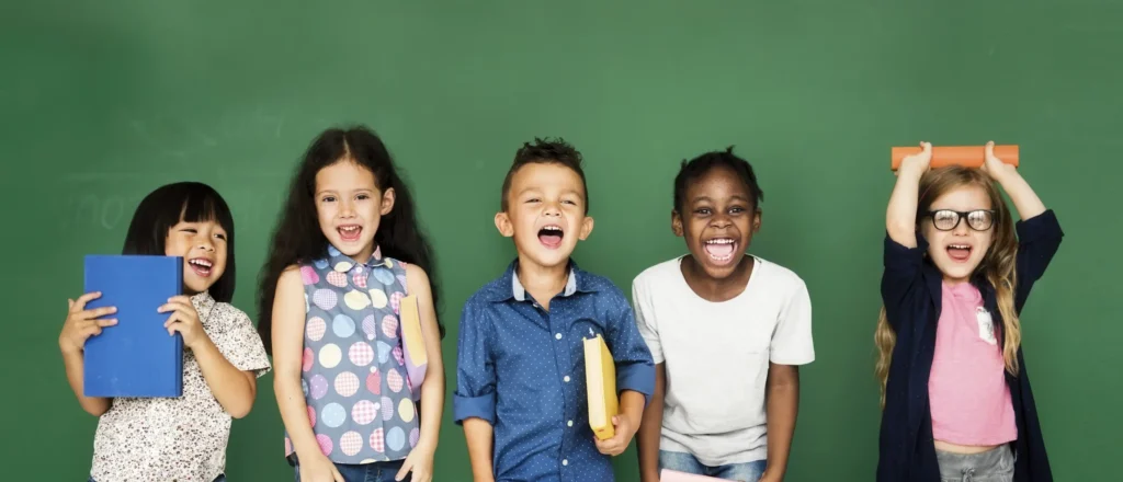 Five cheerful children stand in front of a green chalkboard, smiling and laughing. Some hold books or notebooks, and one child raises an orange book overhead. They appear happy and energetic.