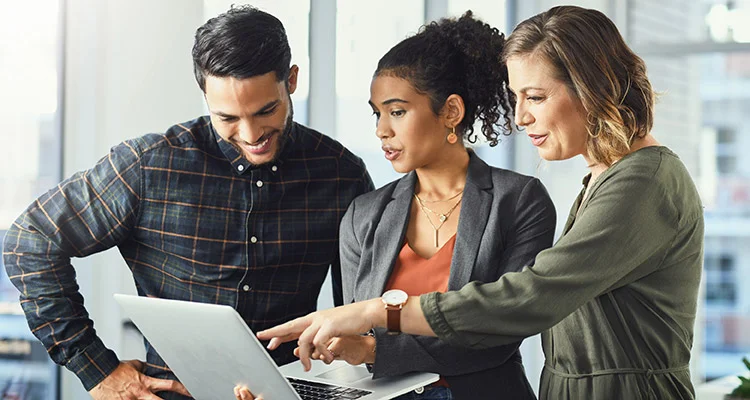Three people stand together in an office, looking at a laptop. One woman points at the screen while the other two, a man and another woman, watch attentively, suggesting a collaborative discussion.