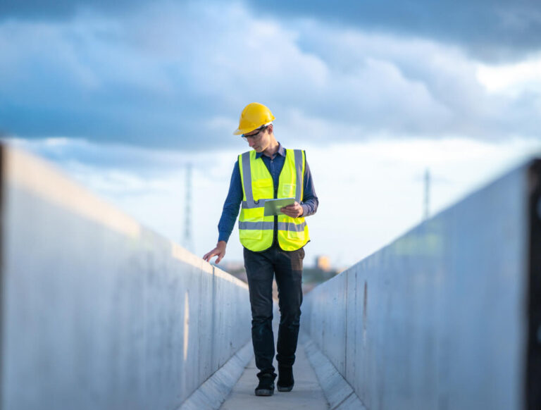 A construction worker wearing a yellow hard hat, safety vest, and blue shirt walks on a concrete path, inspecting the surface while holding a mobile device, with a cloudy sky in the background.