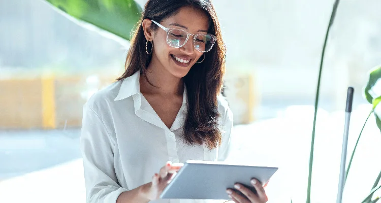 A woman wearing glasses and a white shirt is smiling while using a tablet. She is standing indoors with sunlight streaming in and greenery in the background.