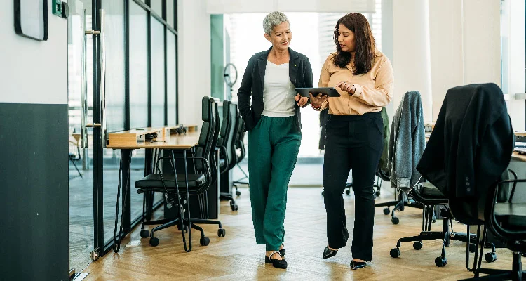 Two women walking through a modern office, one holding a tablet and showing it to the other. They appear engaged in conversation, surrounded by desks, chairs, and large windows.