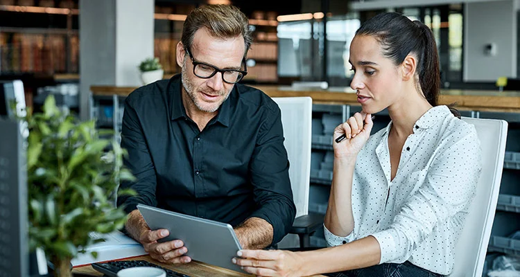 Two colleagues sit at a desk in a modern office, looking at a tablet together. The man wears glasses and a dark shirt, while the woman holds a pen and wears a white blouse with black dots.