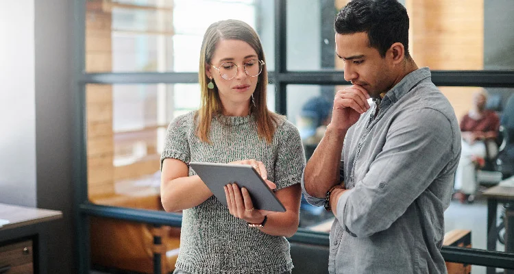 Two colleagues stand in an office, looking at a tablet. The woman is pointing at the screen while the man stands beside her, touching his chin thoughtfully. They appear to be focused and engaged in discussion.