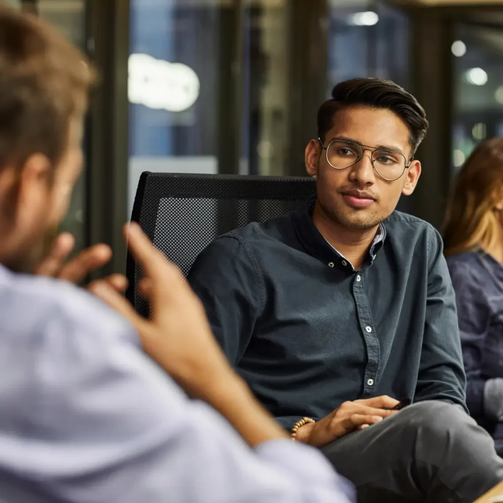A young man wearing glasses and a dark button-down shirt sits and attentively listens to a colleague during an office meeting. Another person is visible in the background, slightly out of focus.