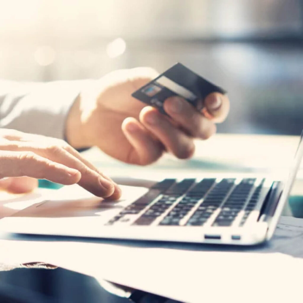 A person holds a credit card in one hand while typing on a laptop keyboard with the other, suggesting online shopping or electronic payment.
