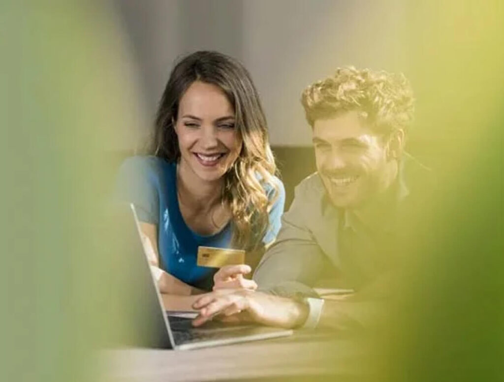 A woman and a man smiling while looking at a laptop together, seen through out-of-focus green foliage in the foreground.