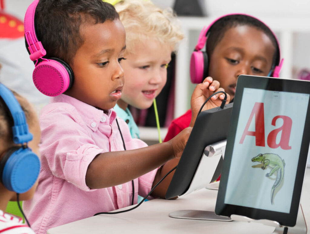 Three young children wearing headphones use tablets in a classroom. The tablet screen in front shows the letter A and a picture of an alligator, suggesting an educational activity.