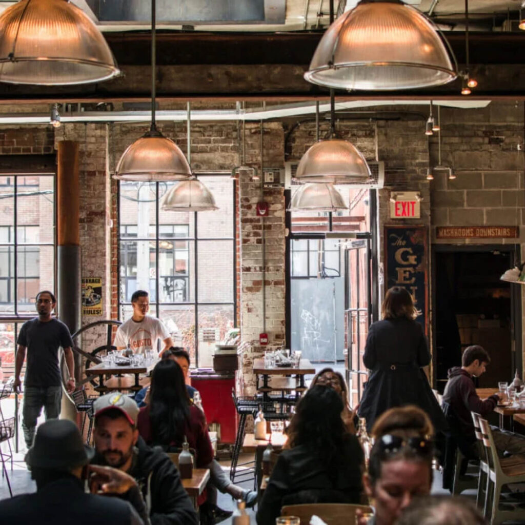 A busy, rustic-style restaurant with exposed brick walls, large windows, industrial hanging lights, and people dining and talking at tables. Two staff members stand near the entrance in the background.