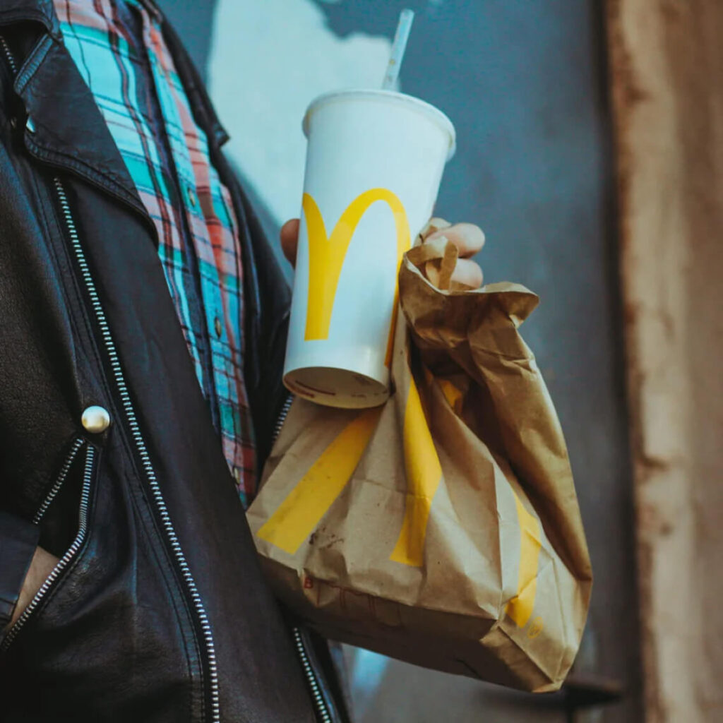 A person wearing a leather jacket and plaid shirt holds a McDonalds paper bag and a McDonalds soft drink cup with a straw.