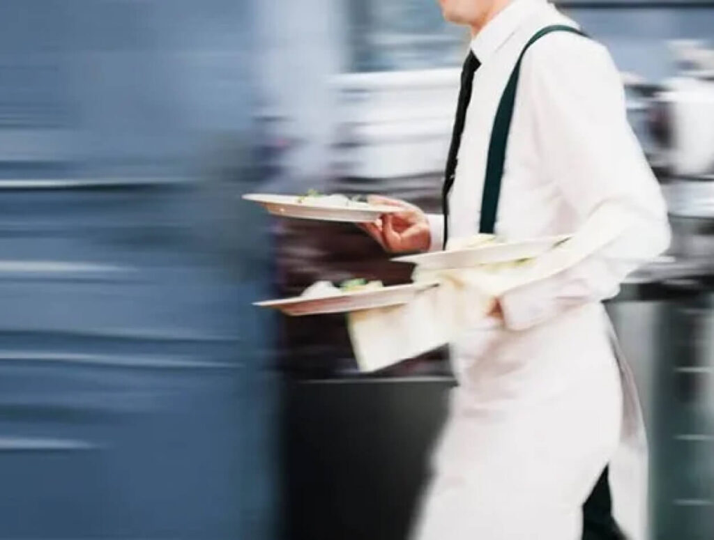 A waiter in a white shirt and apron quickly carries several plates of food in a busy restaurant setting, with a blurred background indicating motion.