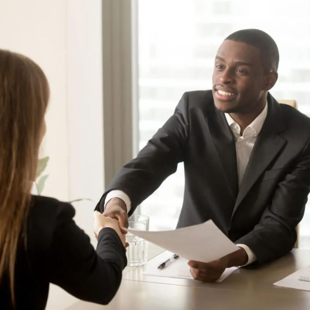 Two people sit together at a desk, looking at papers and shaking hands. They appear focused and engaged in their conversation.