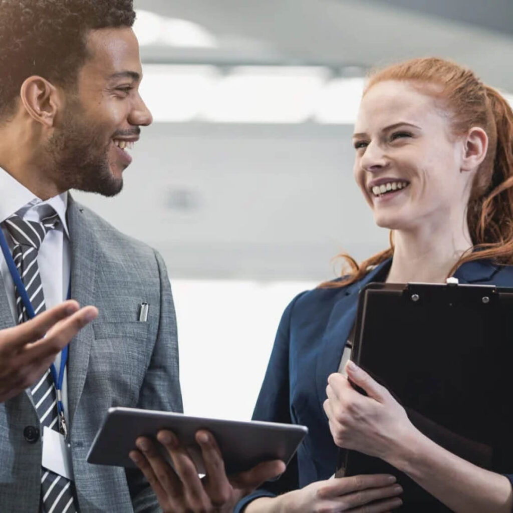 A man in a suit holding a tablet and a woman holding a clipboard are smiling and conversing in a bright office setting.