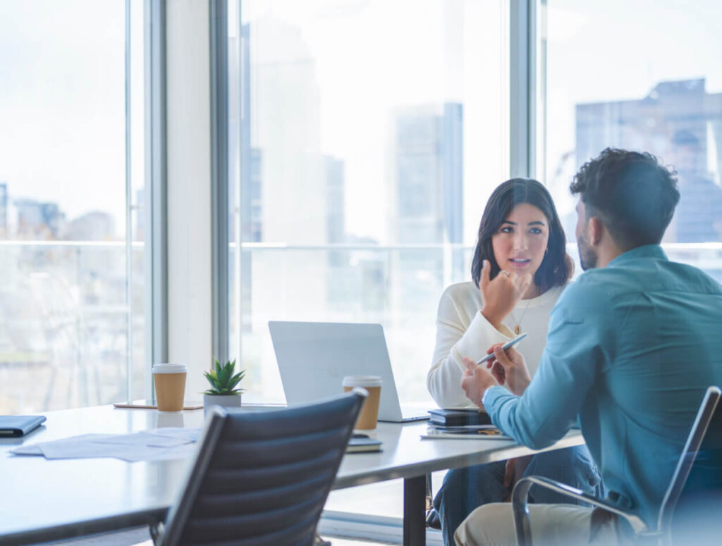 Two people sit across from each other at a conference table in a modern office, engaged in conversation. There are laptops, coffee cups, documents, and a small plant on the table. Large windows show a city view outside.