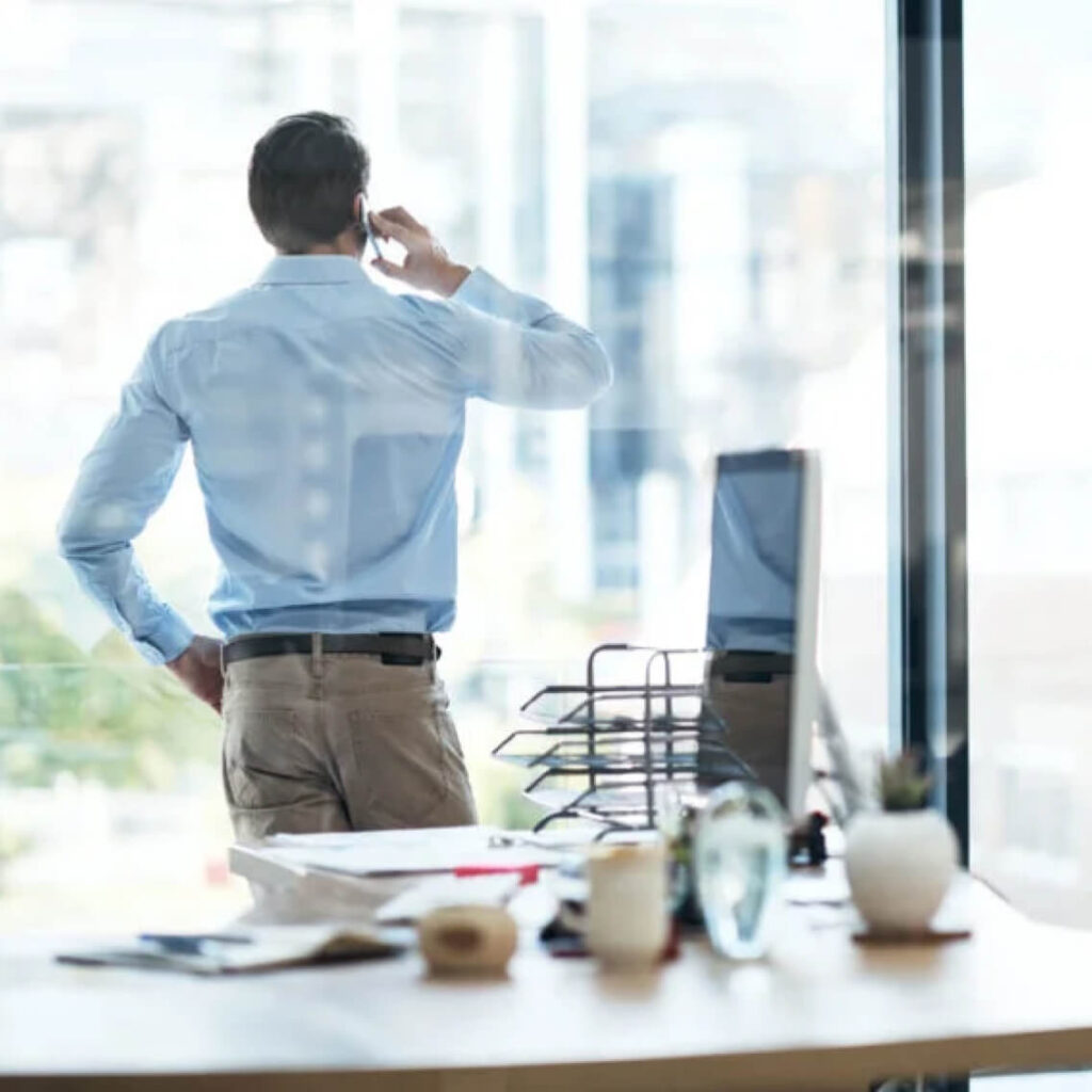 A man in a light blue shirt stands by a large window, talking on a phone. He faces away from the camera. In the foreground, a desk is cluttered with office supplies and potted plants.