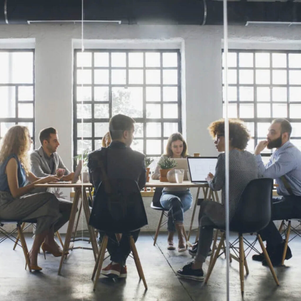 A group of eight people sits around a table in a modern, sunlit office, engaged in discussion. Laptops, papers, and coffee cups are on the table. Large grid windows are in the background.