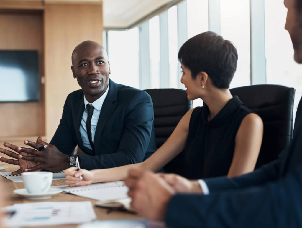 Three professionals in business attire sit at a conference table with notepads and coffee cups, engaged in a friendly discussion in a modern office with large windows.