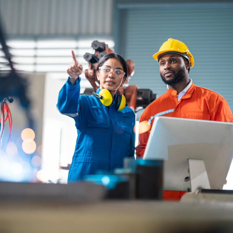 Two engineers in protective gear stand at a workstation in a factory. The woman in blue coveralls points ahead while the man in an orange coverall and yellow hard hat looks on. Industrial equipment is visible around them.