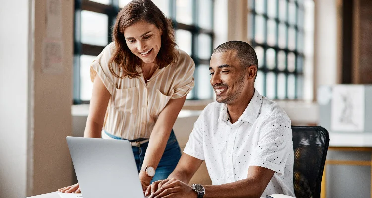 A woman stands next to a man who is sitting at a desk, both smiling and looking at a laptop screen in a bright, modern office setting.