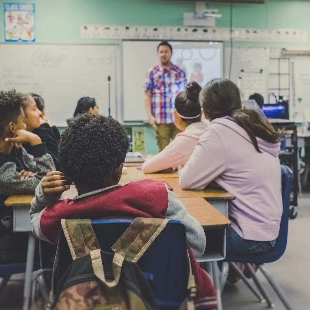 Students sit at desks in a classroom, facing a teacher standing at the front near a projection screen. The teacher appears to be giving a lesson, and students are attentively listening. Classroom walls have posters and whiteboards.